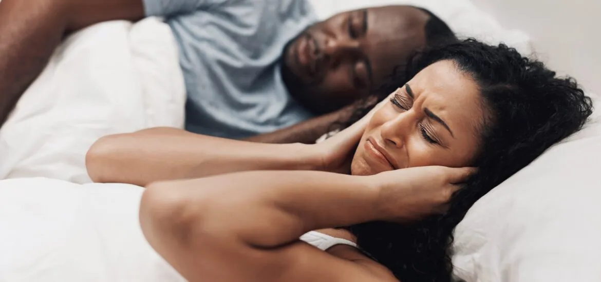 couple in bed, man snoring and disrupting partner who is covering her ears